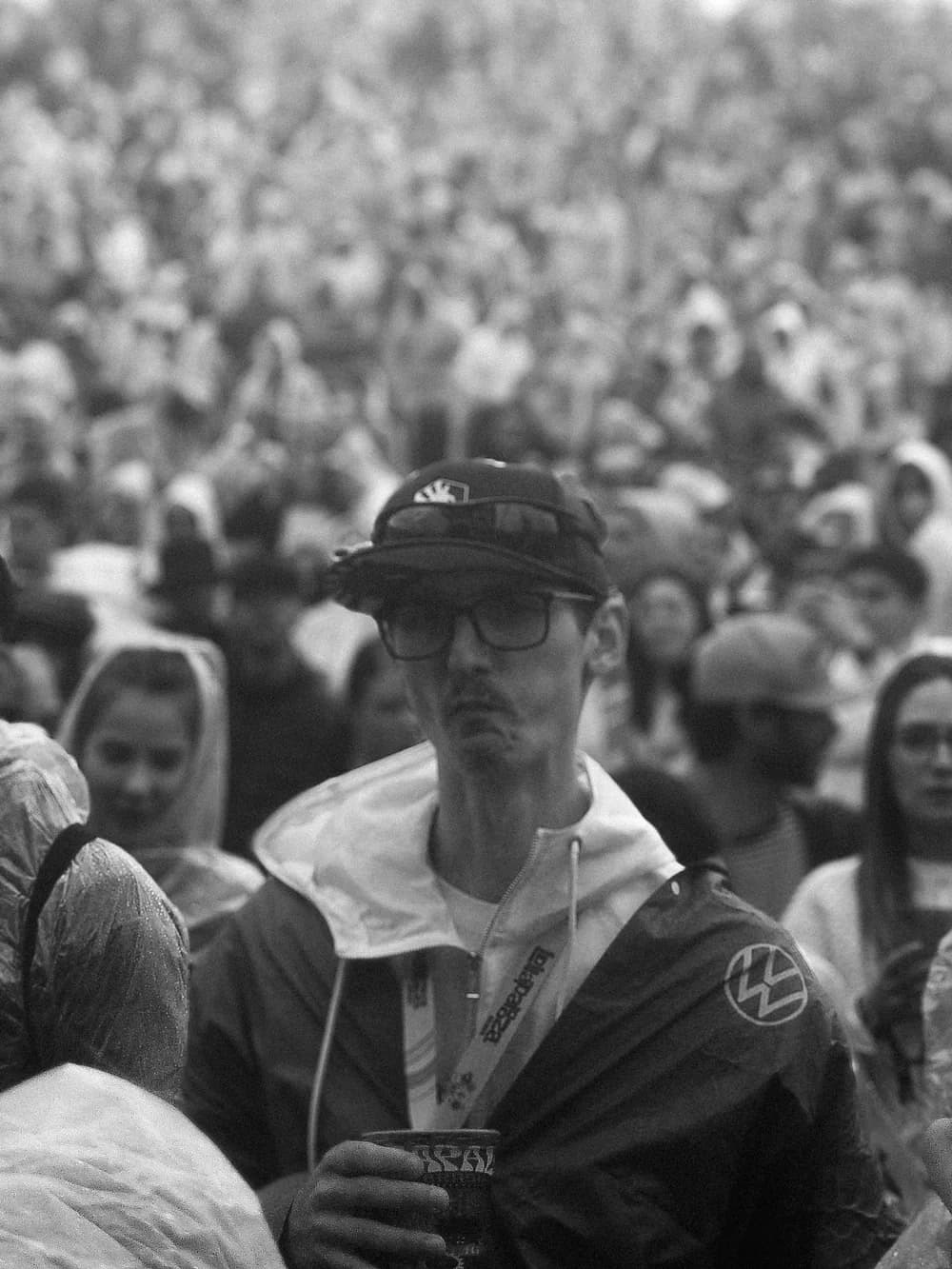 Black and white portrait of a man in a cap, glasses and rain poncho, standing still in a dense festival crowd.