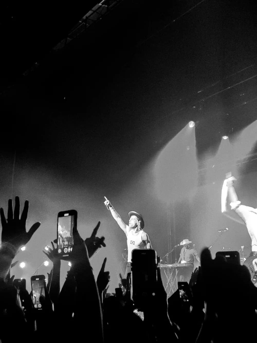 Black and white concert: a singer pointing from a smoky stage while phones and hands rise from the crowd in silhouette.