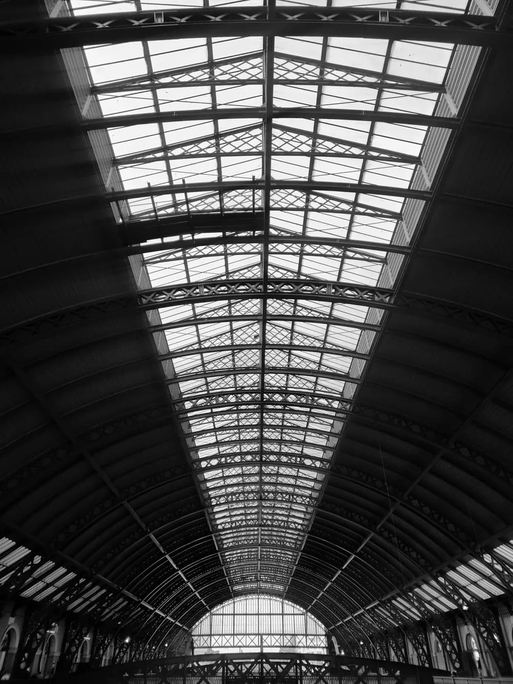 Black and white view up into an iron and glass train station roof, its arched trusses running towards a bright end window.