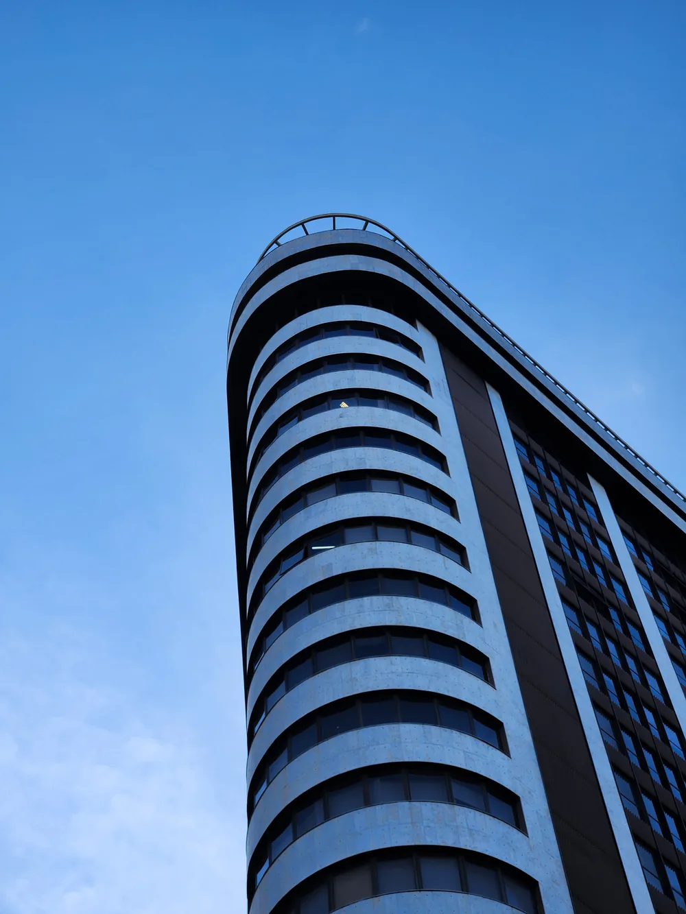 A blue-grey office tower with a rounded corner of curved window bands, seen from below against a clear blue sky.