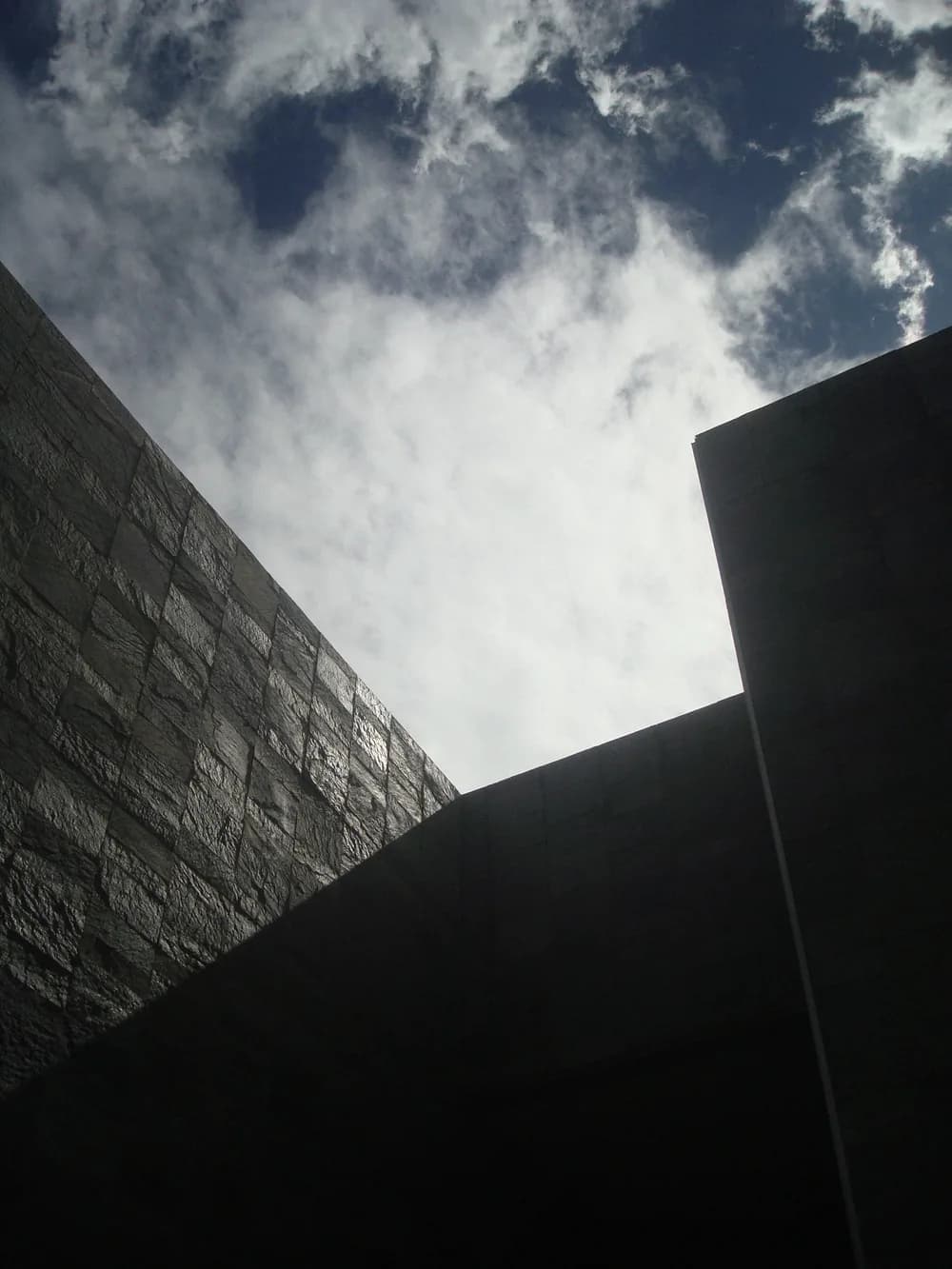 Looking up between dark stone walls at a bright, cloud-streaked sky.