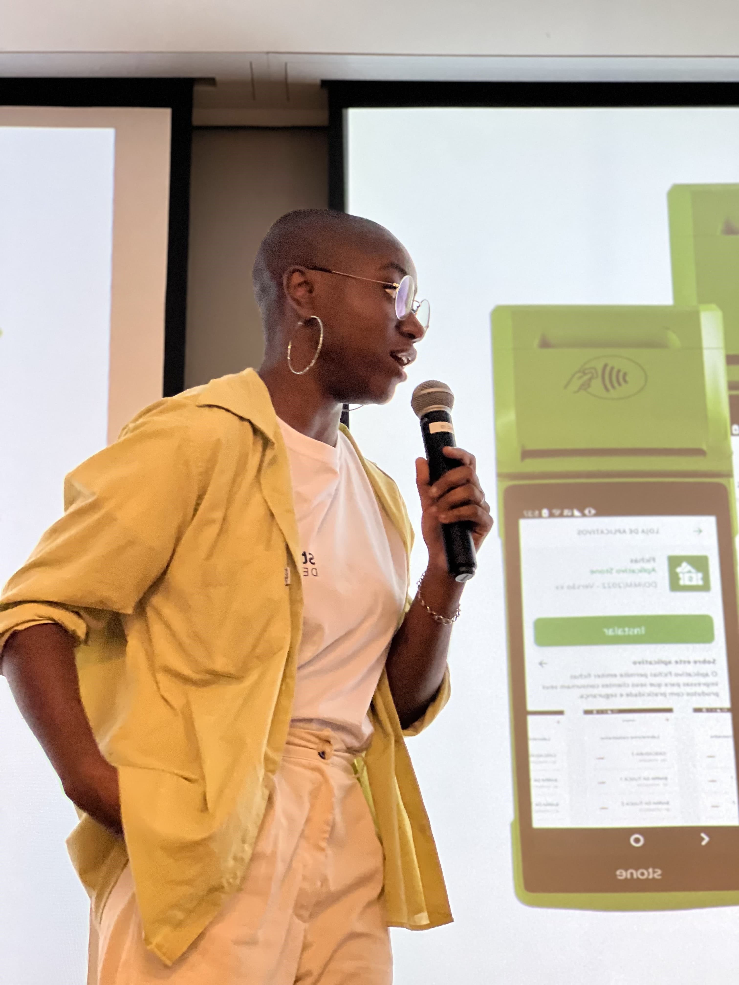 Nicoly Dandara speaking into a microphone beside a projected slide of a Stone payment terminal running the app store.
